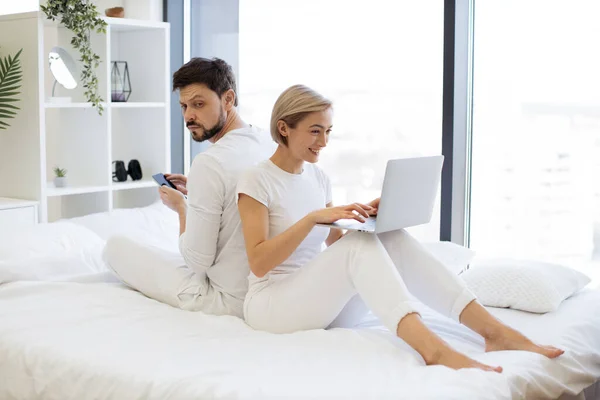 Cheerful amorous people sitting back to back while spending time together in spacious apartment at weekend. Joyful caucasian family scrolling social media on wireless gadgets.