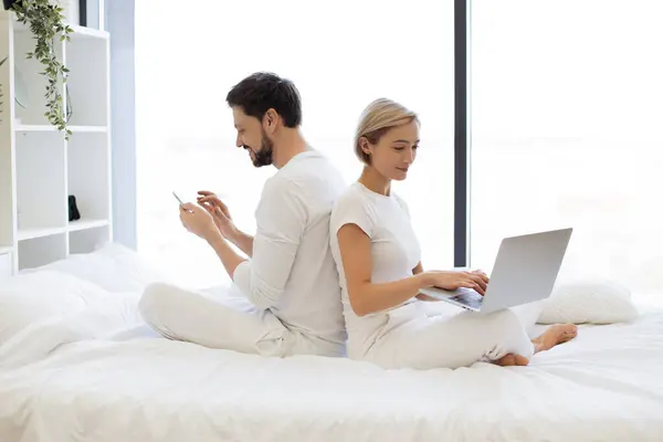 Cheerful amorous people sitting back to back while spending time together in spacious apartment at weekend. Joyful caucasian family scrolling social media on wireless gadgets.