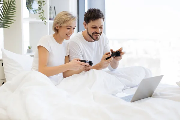 Caucasian couple playing video games while lying under blanket at home. Smiling man and woman have fun competing in laptop game using joysticks.