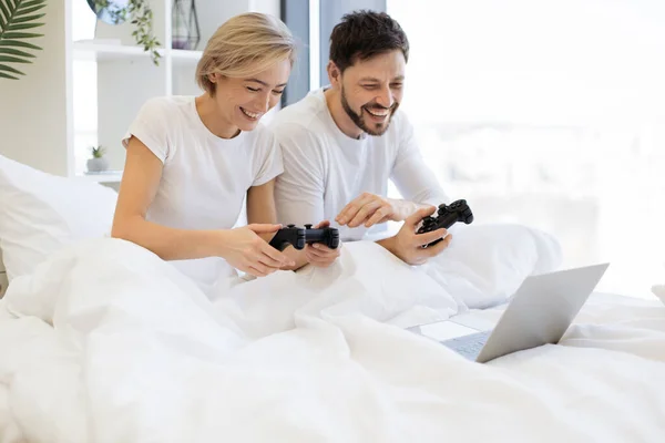 Caucasian couple playing video games while lying under blanket at home. Smiling man and woman have fun competing in laptop game using joysticks.