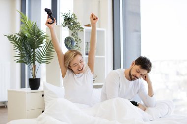 Hüzünlü adam joystick 'i tutuyor ve mutlu karısına bakıyor. Video oyununda zaferi kutlayan elini kaldırıyor. Kafkasyalı aşık çiftin yan görüntüsü yatakta joysticklerle dizüstü bilgisayar oyununda yarışırken iyi eğlenceler..