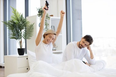 Hüzünlü adam joystick 'i tutuyor ve mutlu karısına bakıyor. Video oyununda zaferi kutlayan elini kaldırıyor. Kafkasyalı aşık çiftin yan görüntüsü yatakta joysticklerle dizüstü bilgisayar oyununda yarışırken iyi eğlenceler..