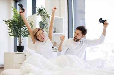 joystick 'le elini kaldıran mutlu kadın video oyununda zaferi kutlayan kocasını izliyor. TV seyreden beyaz bir çiftin yan görüntüsü. Yatakta video oyununda yarışırken eğlenin..