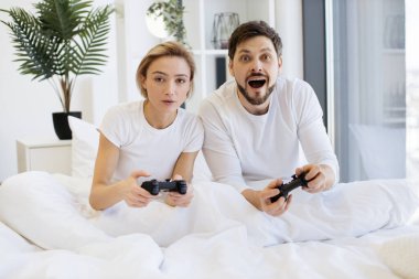 Evde battaniyenin altında yatarken konsantre olmuş beyaz bir çiftin oyun oynamalarının ön görüntüsü. Televizyonda gülümseyen adam ve kadın, joysticklerle video oyununda yarışırken eğleniyor..