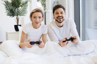 Evde battaniyenin altında yatarken konsantre olmuş beyaz bir çiftin oyun oynamalarının ön görüntüsü. Televizyonda gülümseyen adam ve kadın, joysticklerle video oyununda yarışırken eğleniyor..