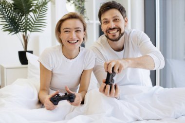 Evde battaniyenin altında yatarken konsantre olmuş beyaz bir çiftin oyun oynamalarının ön görüntüsü. Televizyonda gülümseyen adam ve kadın, joysticklerle video oyununda yarışırken eğleniyor..