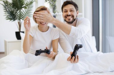 Modern TV 'de joysticklerle video oyunları oynarken yatakta oturan ve karısının gözlerini aldatan genç bir adamın ön görüntüsü. Evlerinde dinlenen evli, beyaz bir çift..