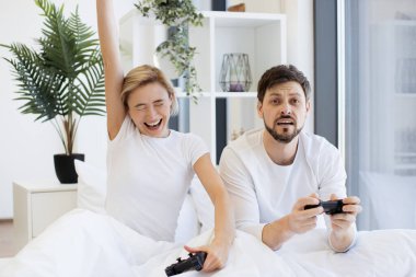 Hüzünlü adam joystick 'i tutuyor ve mutlu karısına bakıyor. Video oyununda zaferi kutlayan elini kaldırıyor. Kafkasyalı aşık çiftin yan görüntüsü yatakta joysticklerle dizüstü bilgisayar oyununda yarışırken iyi eğlenceler..