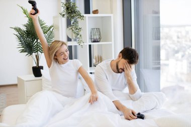 Hüzünlü adam joystick 'i tutuyor ve mutlu karısına bakıyor. Video oyununda zaferi kutlayan elini kaldırıyor. Kafkasyalı aşık çiftin yan görüntüsü yatakta joysticklerle dizüstü bilgisayar oyununda yarışırken iyi eğlenceler..