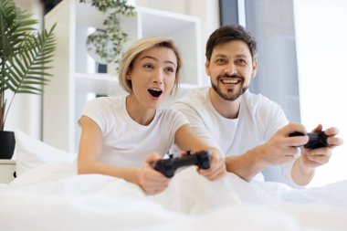 Evde battaniyenin altında yatarken, konsantre olmuş beyaz çiftin video oyunları oynamasını kapat. Televizyonda gülümseyen adam ve kadın, joysticklerle video oyununda yarışırken eğleniyor..