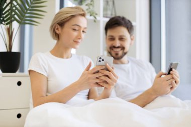 Evde gündüz vakti yumuşak yatakta tembellik ederken cep telefonu kullanan iki kişilik sakin bir beyaz aile. Çevrimiçi makale okuyan kendine güvenen yetişkin erkek ve kadın detaylara dikkat ediyor.