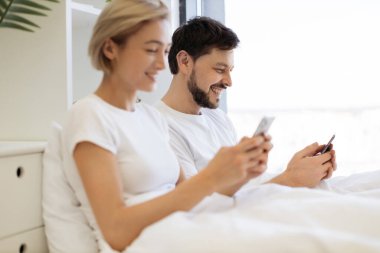 Evde gündüz vakti yumuşak yatakta tembellik ederken cep telefonu kullanan iki kişilik sakin bir beyaz aile. Çevrimiçi makale okuyan kendine güvenen yetişkin erkek ve kadın detaylara dikkat ediyor.