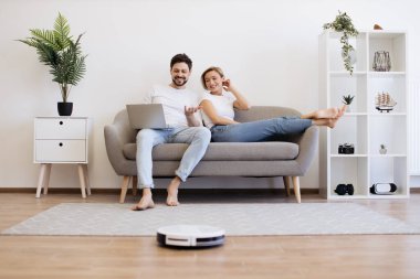 Kafkasyalı genç çift rahat koltukta rahat rahat oturuyor ellerinde taşınabilir laptopları var. Kablosuz robot ahşap zemini süpürürken iki kişilik bir aile film izliyor..