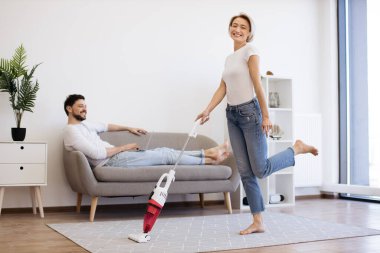 Gündelik giyinmiş neşeli beyaz kadın ev işleri sırasında kablosuz elektrik süpürgesiyle dans ederken kocası kanepeye oturup dizüstü bilgisayarla çalışıyor. Modern genç ailenin yaşam tarzları..