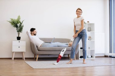 Çekici karısı ev işleri için kablosuz elektrik süpürgesi kullanırken modern dizüstü bilgisayarda çalışan yakışıklı beyaz bir adam. Gündelik giysiler içinde genç ve güzel bir çift boş zamanlarında modern aletlerin tadını çıkarıyor..