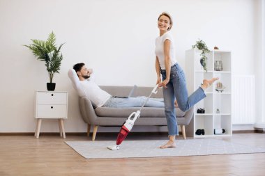 Gündelik giyinmiş neşeli beyaz kadın ev işleri sırasında kablosuz elektrik süpürgesiyle dans ederken kocası kanepeye oturup dizüstü bilgisayarla çalışıyor. Modern genç ailenin yaşam tarzları..