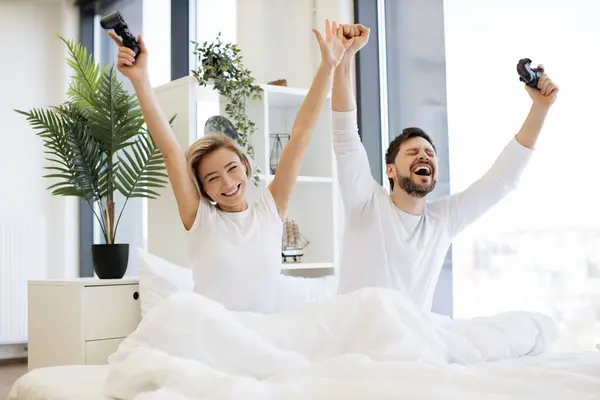 Happy woman with raising hands with joystick looking at husband celebrating victory in video game. Side view of caucasian couple in love looking at tv have fun competing in video game on bed.