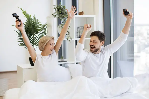 Happy woman with raising hands with joystick looking at husband celebrating victory in video game. Side view of caucasian couple in love looking at tv have fun competing in video game on bed.