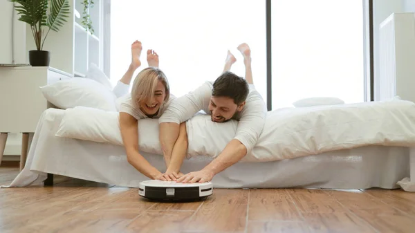 Smiling wife and husband having fun playing with smart cleaning robot while resting on bed at home. Family man and woman creating love bond by spending time together while spending time together.