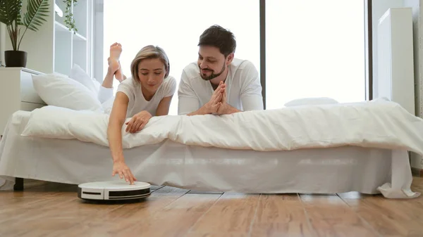 Close up view of joyful woman and man activating robotic cleaner while resting on bed in apartment. Delighted wife and husband following rules of using smart electrical appliance at home.