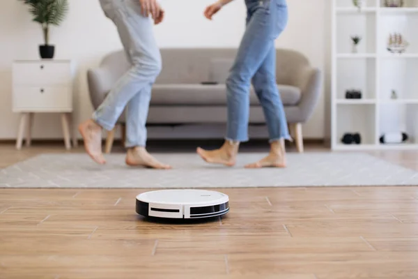 Close up view of modern cleaning robot vacuuming bare laminate floor with cropped view of male and female in background. Automatic gadget dusting off surface while wife and husband dancing for music.