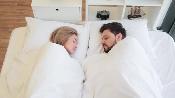 Top view of loving caucasian couple sleeping face to face on soft white bed in modern apartment. Relaxed pretty woman and handsome bearded man having peaceful daytime rest in bright bedroom.