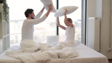 Genç tutkulu beyaz çift yastıklarla kavga ediyor, panoramik pencereli parlak yatak odasında uyandıktan sonra eğleniyorlar..