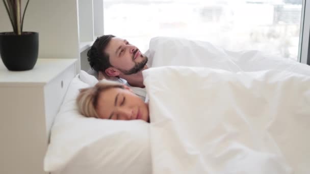 Smiling bearded husband relaxing on bed with pillow under head while sleeping wife resting on background. Cheerful caucasian man in mature relationship relishing full life.