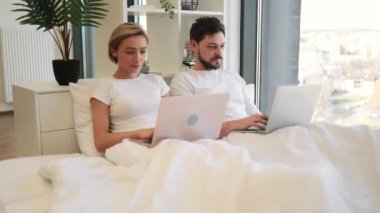Yanındaki portatif bilgisayarda yazı yazmakla meşgul olan beyaz bir erkek dizüstü bilgisayarla yatakta sayfaları karıştırıyor. İlişkilerde olgun insanlar evde birlikte vakit geçirirler..