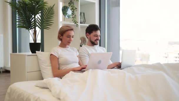 家で毛布の下に横たわっている間 現代のガジェットに取り組むのが大好きの慎重なカップルの側面 笑顔の男は ノートパソコンを使用してソーシャルアップデートを読み 女性はオンラインでショッピング — ストック動画