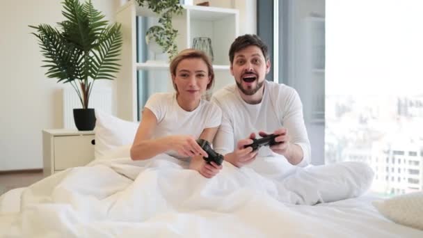 悲しい男はジョイスティックを持って 幸せな妻がビデオゲームで勝利を祝う手を挙げているのを見ています 愛の慎重なカップルの側面のビューは ベッドのジョイスティックを使用してラップトップゲームで楽しい競争を持っています — ストック動画
