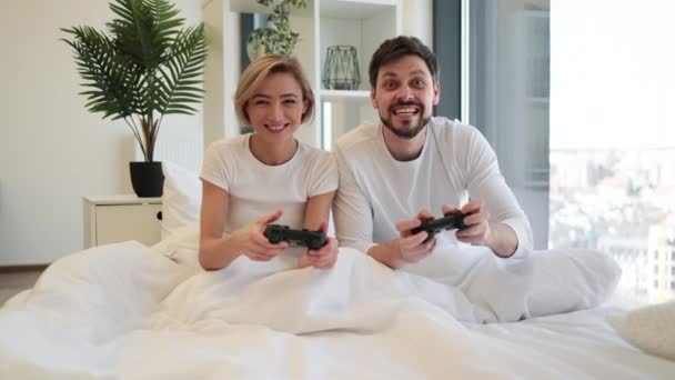 ジョイスティックを持って幸せな夫を見ている悲しい女性は ビデオゲームで勝利を祝う手を挙げました 愛の慎重なカップルの側面のビューは ベッドのジョイスティックを使用してラップトップゲームで楽しい競争を持っています — ストック動画