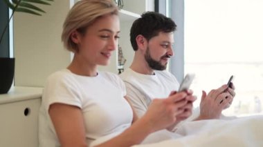 Evde gündüz vakti yumuşak yatakta tembellik ederken cep telefonu kullanan iki kişilik sakin bir beyaz aile. Çevrimiçi makale okuyan kendine güvenen yetişkin erkek ve kadın detaylara dikkat ediyor.