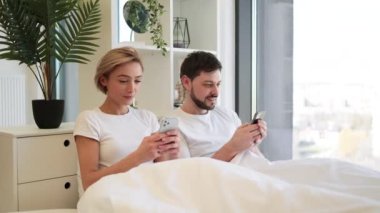 Kafkasyalı mutlu bir çift cep telefonlarını elinde tutarken tüm sabah yumuşak yataklarında uzanıyorlar. Çekici erkek ve kadın dijital aygıtlarda mobil uygulama kullanarak internette geziniyorlar.