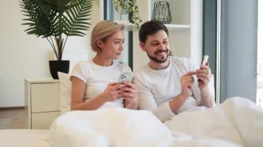 Evde gündüz vakti yumuşak yatakta tembellik ederken cep telefonu kullanan iki kişilik sakin bir beyaz aile. Çevrimiçi makale okuyan kendine güvenen yetişkin erkek ve kadın detaylara dikkat ediyor.