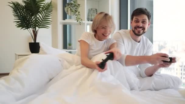 ビデオゲームで勝利を祝う夫を見てジョイスティックで手を挙げた幸せな女性 テレビを見るのが大好きの白人カップルのサイドビューは ベッドのビデオゲームで楽しい競争を持っています — ストック動画