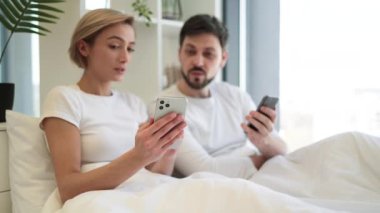 Kafkasyalı mutlu bir çift cep telefonlarını elinde tutarken tüm sabah yumuşak yataklarında uzanıyorlar. Çekici erkek ve kadın dijital aygıtlarda mobil uygulama kullanarak internette geziniyorlar.