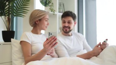 Bütün sabah evde yumuşak bir yatakta uzanırken mutlu beyaz bir çiftin akıllı telefonunu tutmaya odaklan. Çekici erkek ve kadın dijital aygıtlarda mobil uygulama kullanarak internette geziniyorlar.