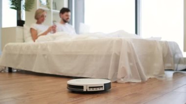 Genç çok ırklı çift dizüstü bilgisayarda video izlerken modern robot süpürgesinin yatak odasındaki ahşap zemini temizleyişinin seçici görüntüsü. Genç bir aile, elektrikli alet kullanarak boş zaman tasarrufu yapıyor..