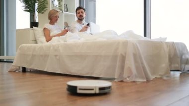 Kafkasyalı sevimli çift rahat bir yatakta uzanıyorlar ve robotlar evdeki ahşap zemini süpürürken modern laptop kullanıyorlar. Modern aygıtlarla ev işleri.