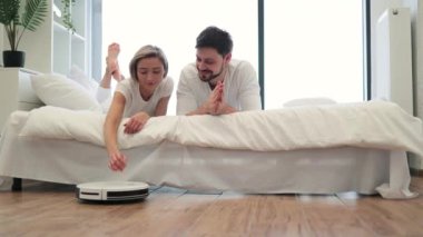 Neşeli kadın ve adamın dairede yatakta dinlenirken robot temizleyiciyi aktif hale getirmesi. Evde akıllı elektrikli alet kullanma kurallarına uyan mutlu bir eş ve koca..
