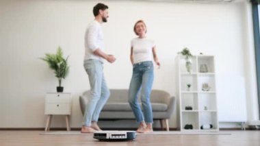 Evde dans ederken yerde çalışan robot temizleyicisiyle dans eden sıradan giysili neşeli bayan. Mutlu beyaz aile babası ve karısı evde bakım yaparken eğleniyorlar..