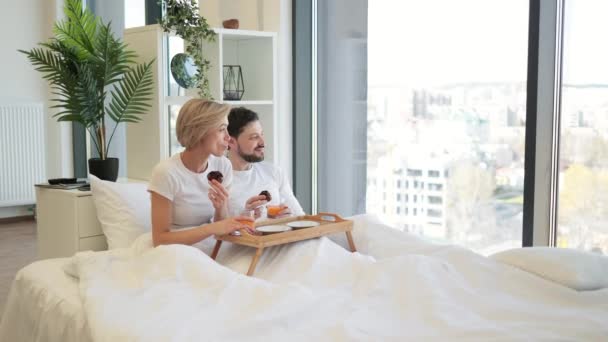 白人家庭的男人和女人躺在床上吃松饼 喝橙汁 快乐的已婚夫妇看着全景窗口 通过日常护理传递着真爱的信息 — 图库视频影像