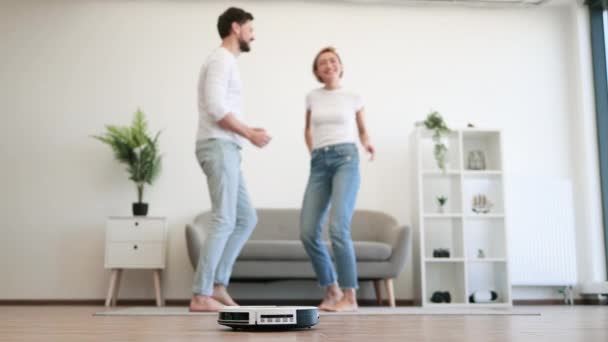 ホームダンスパーティー中に夫と踊るカジュアルな服の陽気な女性は 床の作業ロボット掃除機で 幸せな白人家族の男と彼の妻は ホームケアをしているときに楽しんでいます — ストック動画