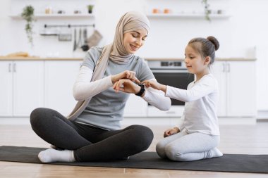 Kızı olan Müslüman bir kadın, mutfakta yoga minderinde otururken spor nöbetlerinde antrenman yapıyor. Anne ve kız arkadaşı spor takip cihazını kullanarak ev eğitiminden faydalanıyorlar..