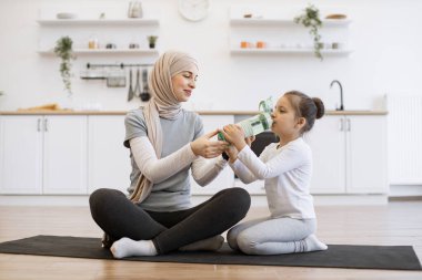 Aktif giyimli güzel Müslüman kadın elinde bir şişe su tutarken tatlı kızı lastik minderde otururken içki içiyor. Sağlıklı kadın ebeveyn ve küçük çocuk jimnastik sırasında sıvı kaybını telafi ediyorlar..