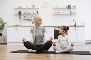 Yoga pozisyonunda oturan rahat Müslüman bir kadının modern dairede lastik minder üzerinde küçük bir çocukla tam boy görüntüsü. Gyan Mudra 'da parmaklarıyla egzersiz yapan sağlıklı bir anne ve birbirine bakan bir kız.