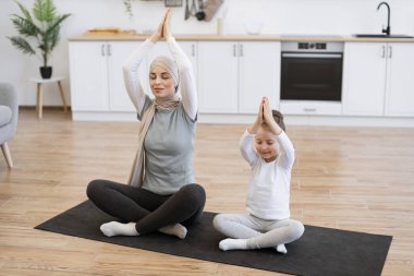 Spor kıyafetleri içinde küçük kızı olan Müslüman bir kadın ve spor salonunda bacak bacak bacak üstüne atarak meditasyon yaparken avuçlarını kapalı tutuyor. Birlikte yoga yapan sağlıklı bir aile.