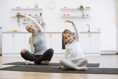 Yoga minderindeki sıska müslüman kadın küçük bir kızla çalışırken kollarını esnetiyor. Spor giyim sektöründe genç bir ebeveyn ve kız spor yaparken eğleniyor..