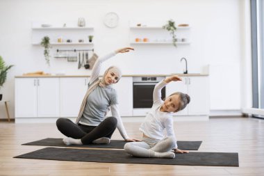 Yoga minderindeki sıska müslüman kadın küçük bir kızla çalışırken kollarını esnetiyor. Spor giyim sektöründe genç bir ebeveyn ve kız spor yaparken eğleniyor..